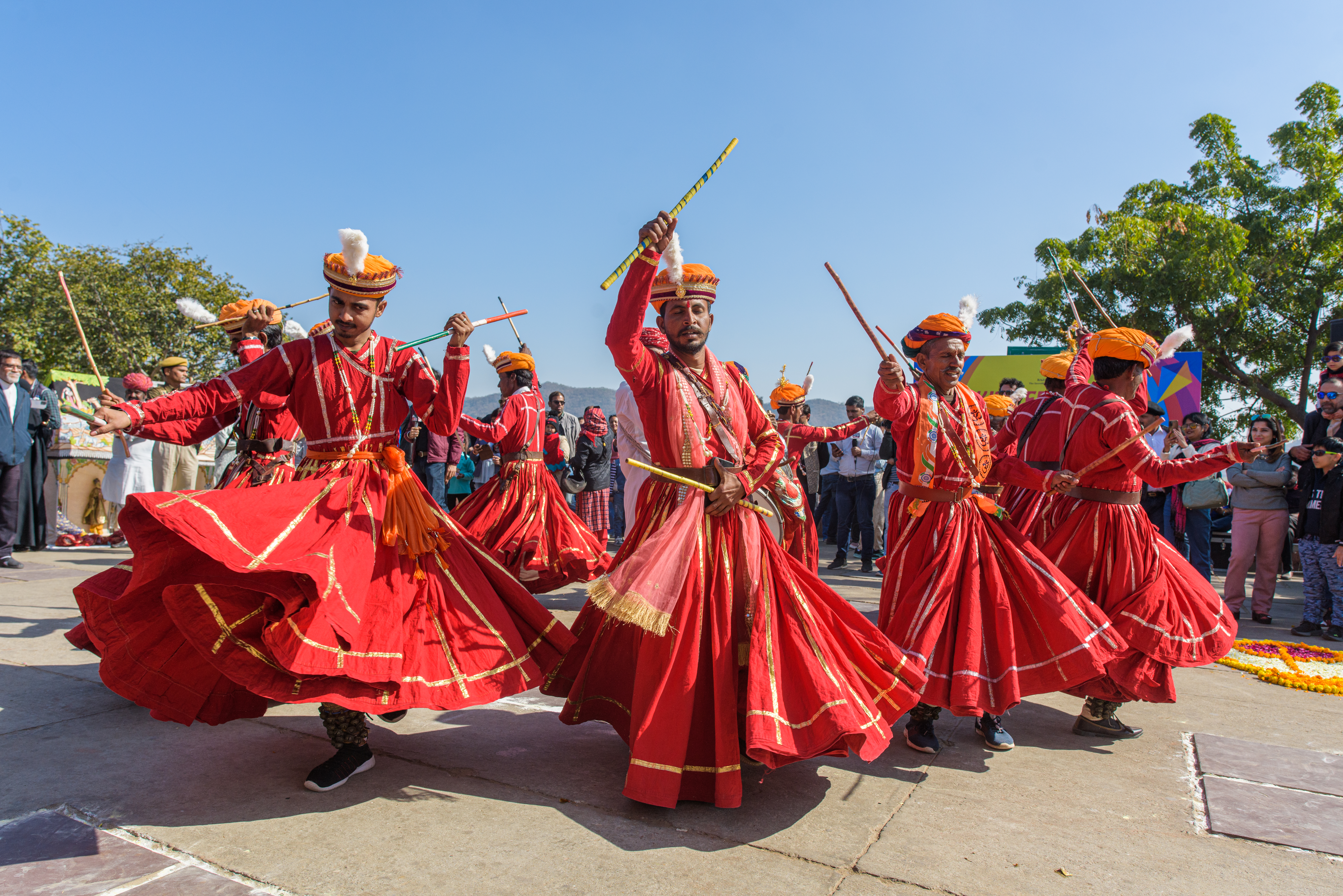 A group of performers dressed in vibrant red traditional attire with orange turbans are dancing energetically in an outdoor setting. They are holding colourful sticks, suggesting a folk dance, while their flowing garments create dynamic movement. The background shows a clear blue sky, trees, and a crowd of spectators watching the performance.