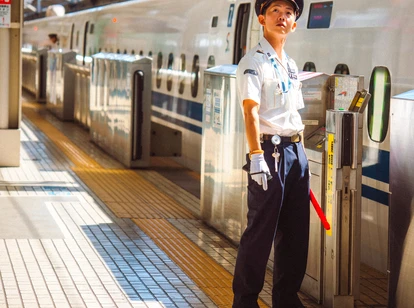 A uniformed railway staff member in Japan stands on a tiled platform beside a sleek white high-speed train. The person is wearing a white shirt, dark trousers, gloves, and a peaked cap, holding a red baton in one hand. The scene is brightly lit, with sunlight casting shadows across the platform, and the train doors are aligned with safety barriers.